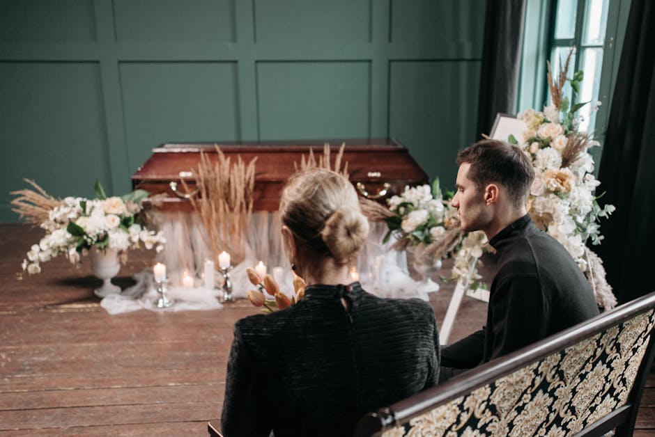 A floral arrangement featuring soft pink and white roses, peonies, and delicate greenery, displayed in a white vase on the floor of a funeral setting. The arrangement is complemented by pampas grass a