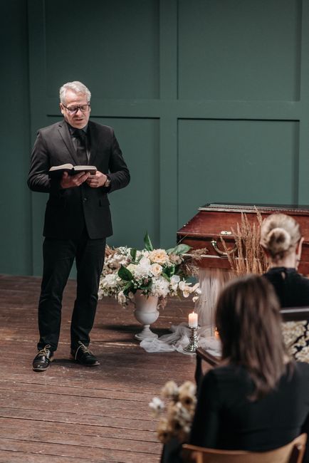 A man wearing glasses and a dark suit stands holding an open book, possibly reading a speech, in front of a floral arrangement of blush pink, white, and green flowers placed in a white vase on a woode