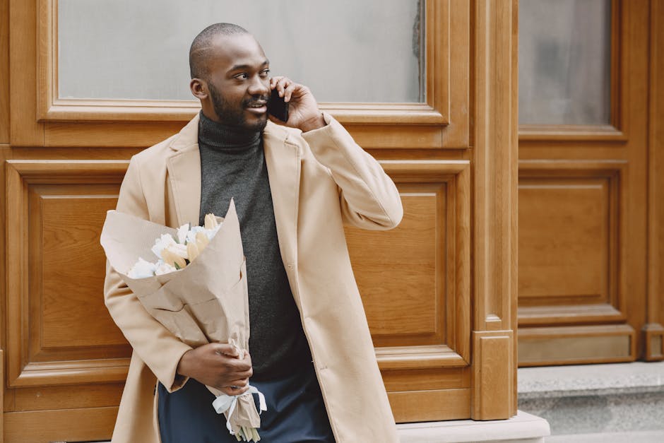 A smiling man with dark skin, a trimmed beard, and short hair, standing outdoors against a wooden-paneled building, is holding a bouquet of fresh white and pale yellow flowers wrapped in brown paper i