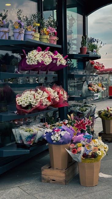 A display of freshly arranged flower bouquets on black metal shelving outside a florist shop. The arrangements include white, pink, and purple flowers such as lilies, chrysanthemums, and roses, wrappe
