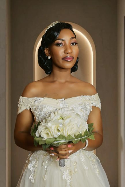 The image features a bride holding a floral bouquet in front of her, dressed in an elegant off-the-shoulder ivory wedding gown adorned with intricate lace and beadwork. Her dark hair is styled in soft
