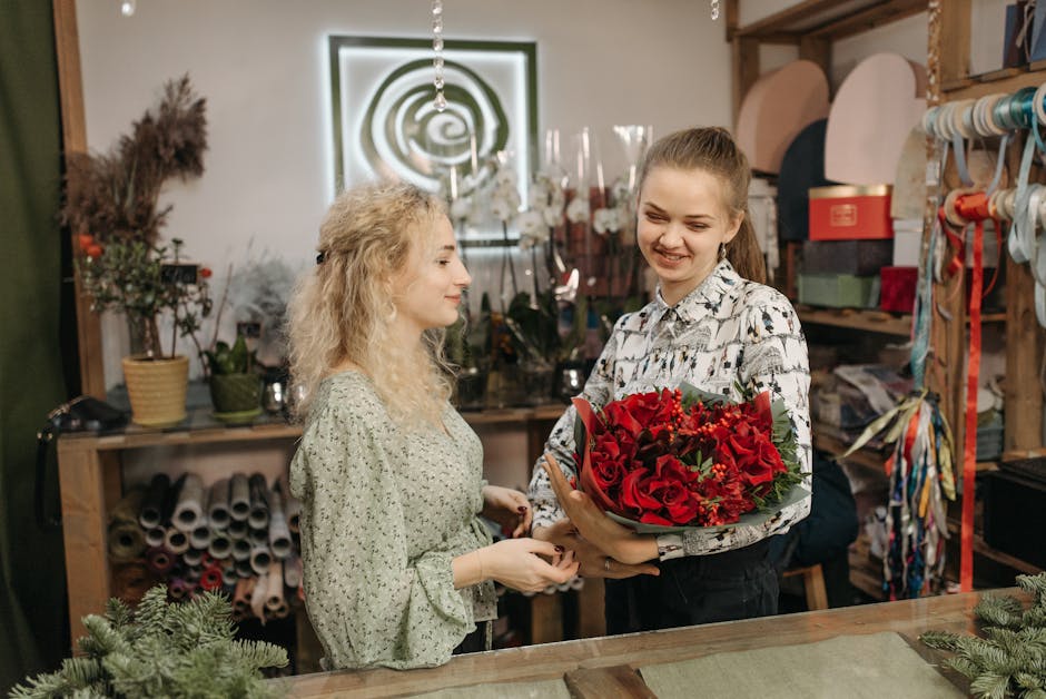 A young woman with a cheerful expression and light brown hair tied back, is holding a vibrant bouquet of fresh red roses, deep red hypericum berries, and green foliage, wrapped in brown paper and secu